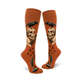 Pair of rust-colored knee-high socks. Design includes a raven perched on a skull above an antique book, quill pen, inkwell, and flying birds.