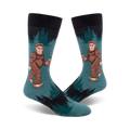 A pair of teal crew socks with a Bigfoot design holding a mug. Black sock top and dark green forest design in the background.