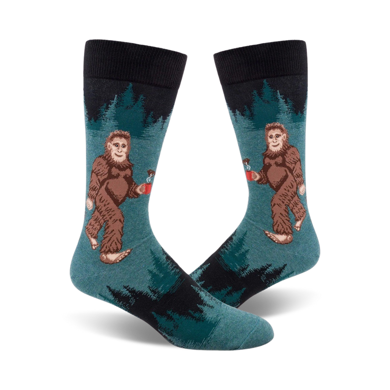 A pair of teal crew socks with a Bigfoot design holding a mug. Black sock top and dark green forest design in the background.