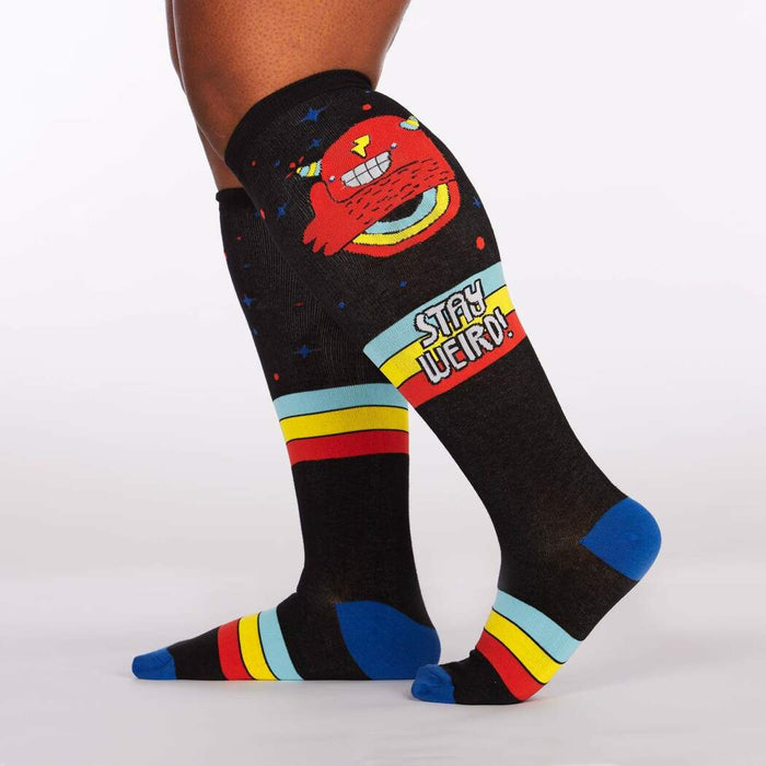 A pair of black knee-high socks with a colorful pattern of monsters and rainbows. The socks have a blue toe and heel, and a red, yellow, and blue striped top. The socks are shown on a plus-size model.