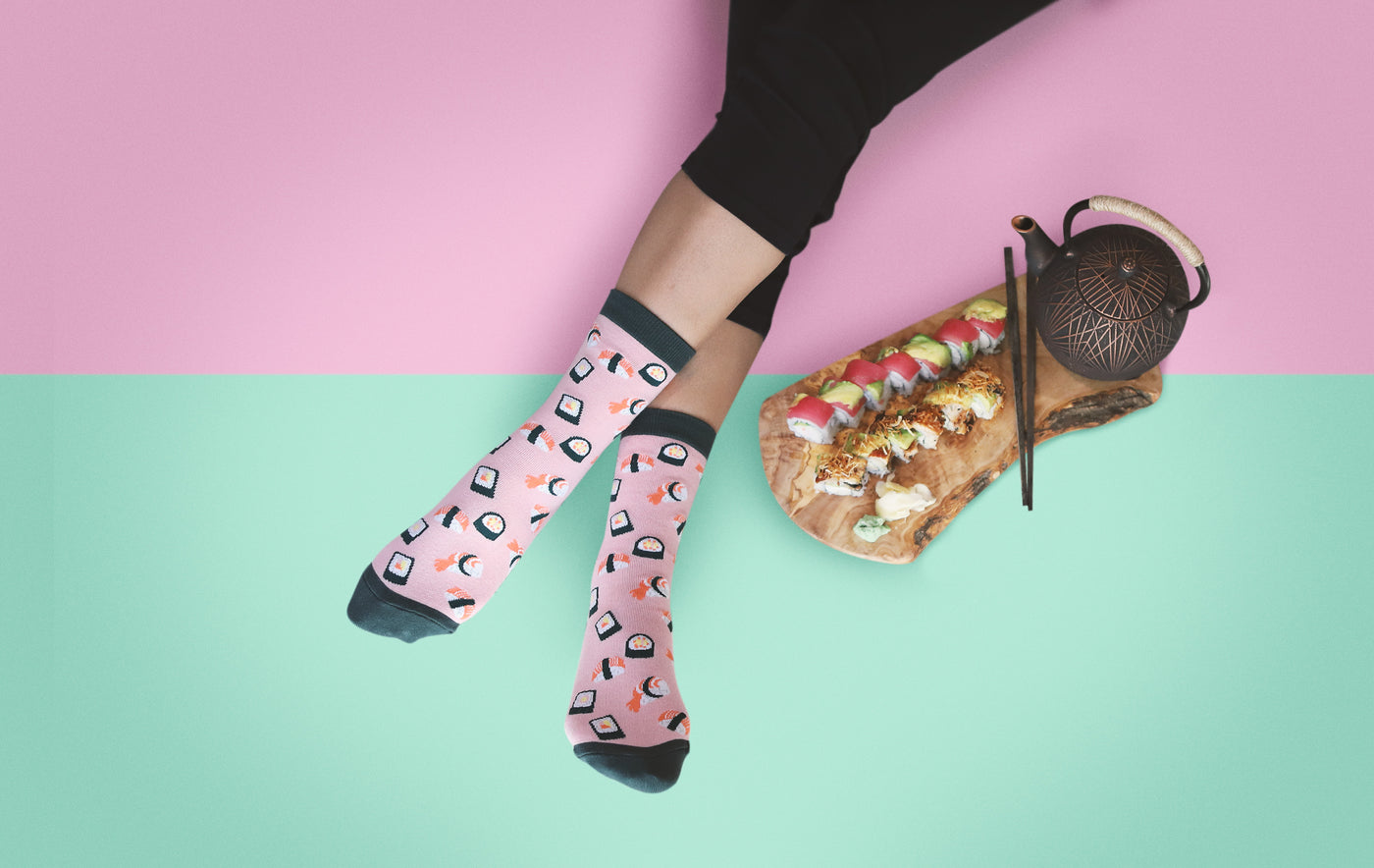 [lf] Legs in pink sushi-patterned socks relax beside a sushi platter and teapot on a pink and mint background.