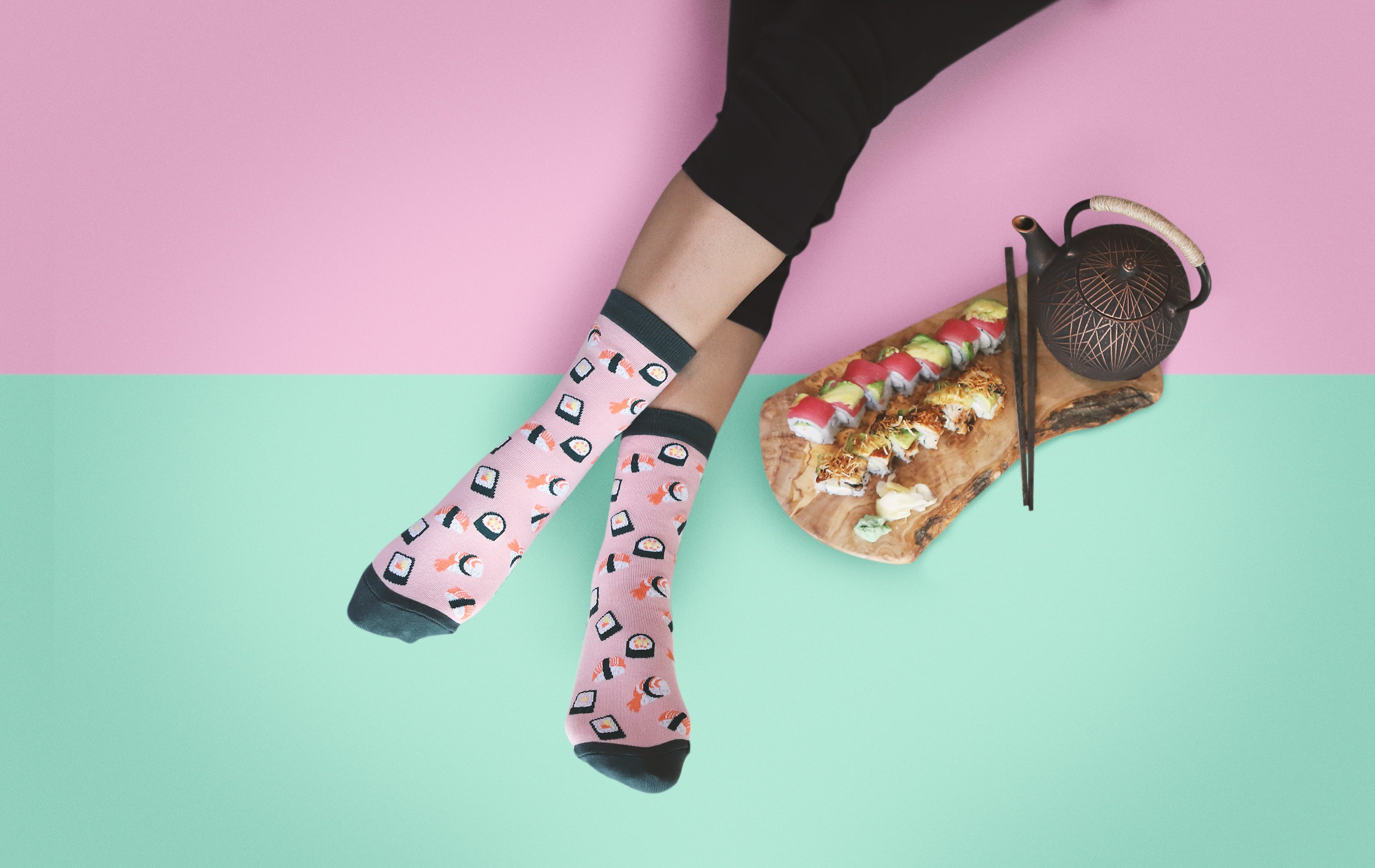[lf] Legs in pink sushi-patterned socks relax beside a sushi platter and teapot on a pink and mint background.