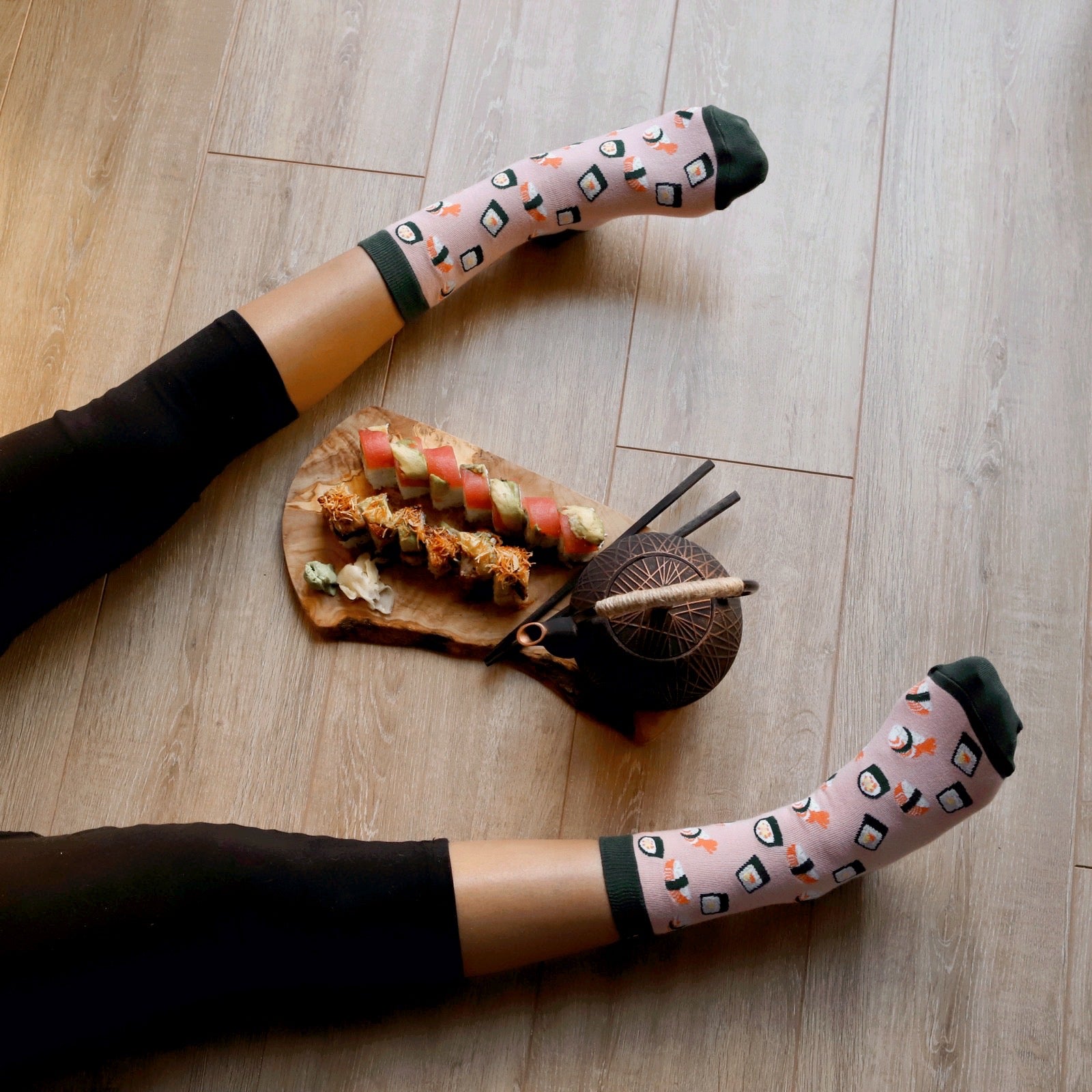 [lf] Legs in black leggings and pink sushi-patterned socks relax on a wood floor with sushi and a teapot.