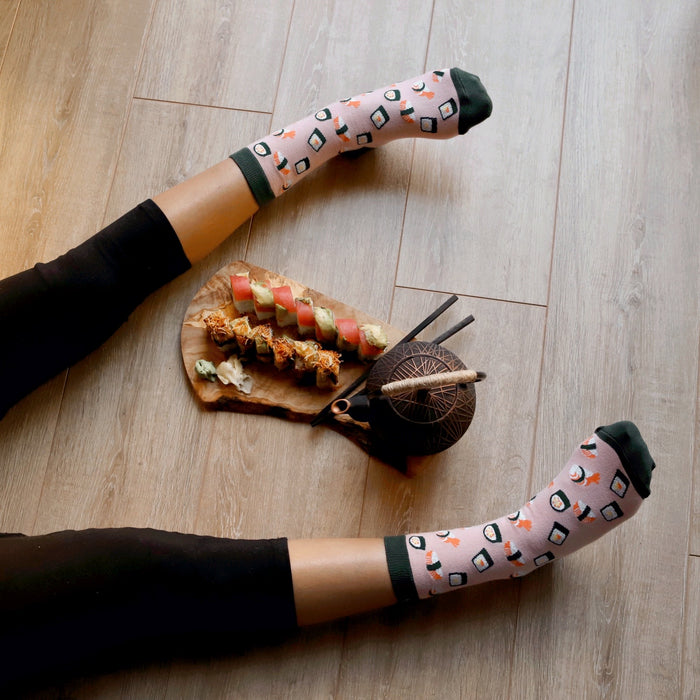 A person is lying on the floor with their feet in the air. They are wearing pink socks with a sushi pattern. There is a wooden board on the floor with a variety of sushi rolls on it. There is also a cast iron teapot on the floor.