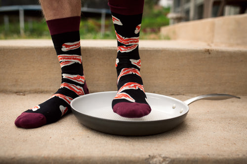 [lf] Person wearing black and red bacon-patterned socks stands with one foot in a frying pan.