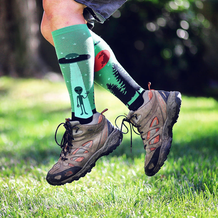 A person is jumping in the air while wearing green socks with an alien and spaceship pattern, dark colored pants, and brown hiking boots. The background is green grass with blurred trees in the distance.