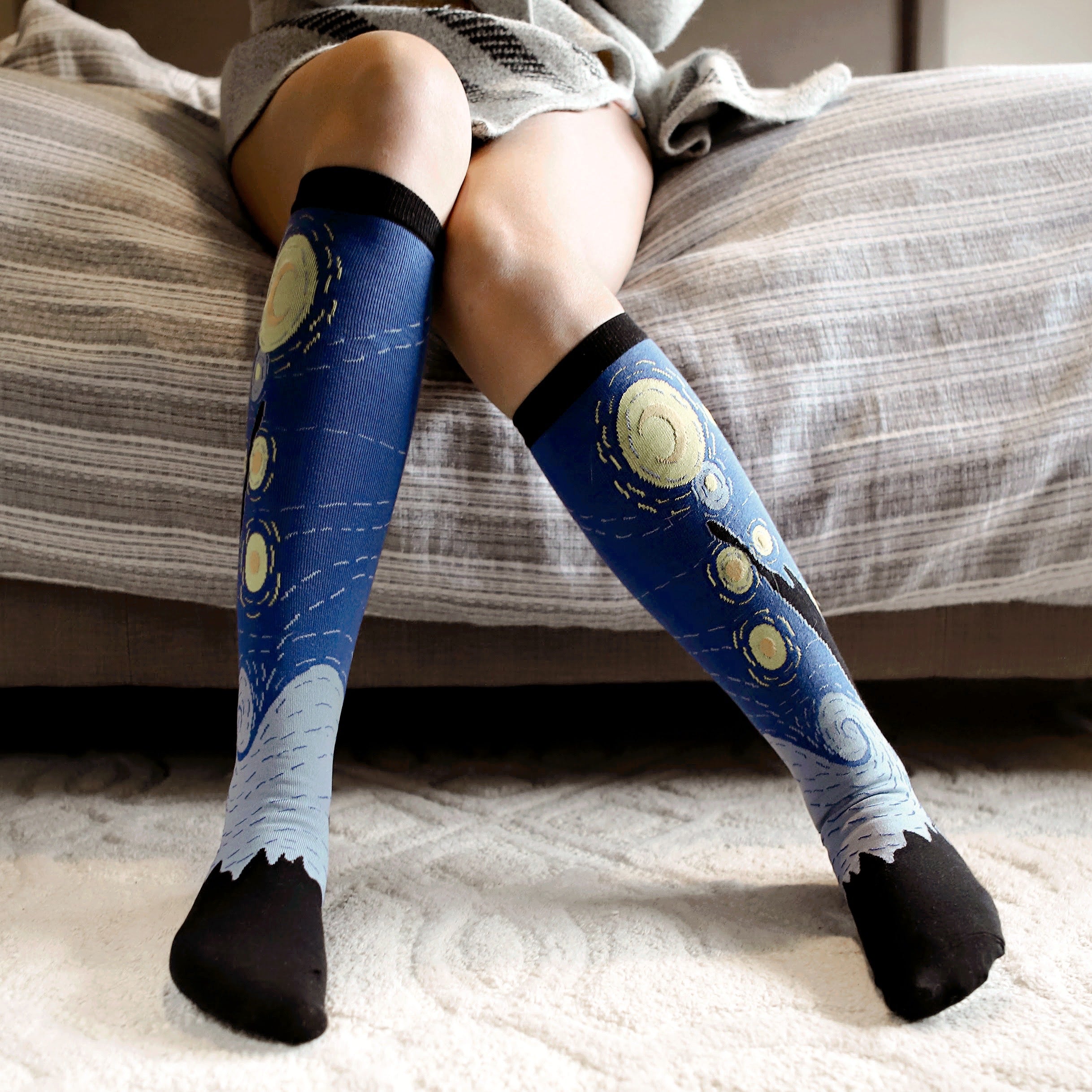 [lf] Woman sitting on bed, wearing Starry Night socks with blue, yellow, and black patterns.