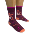 Person wearing dark burgundy socks featuring a white cat tangled in yarn and the phrase Fluff around & find out in orange.