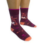 Person wearing dark burgundy socks featuring a white cat tangled in yarn and the phrase Fluff around & find out in orange.