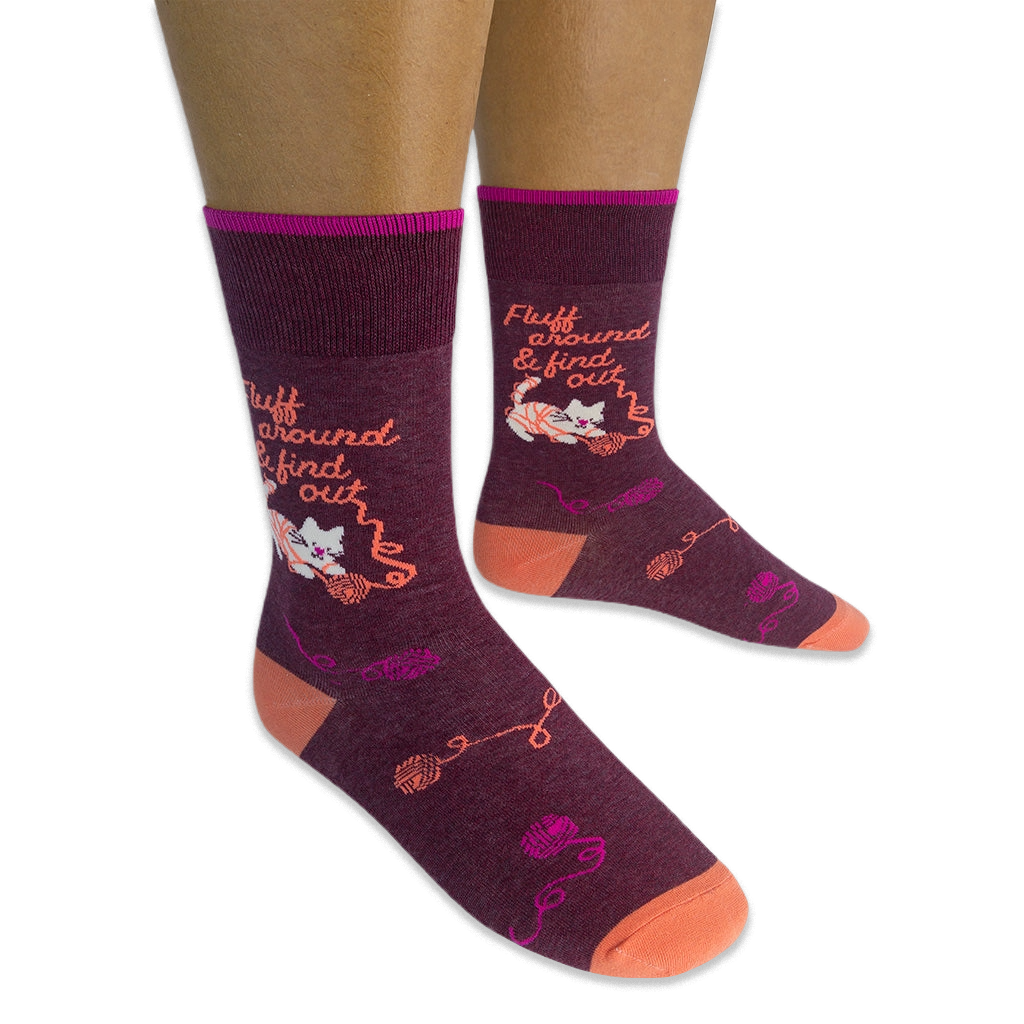 Person wearing dark burgundy socks featuring a white cat tangled in yarn and the phrase Fluff around & find out in orange.