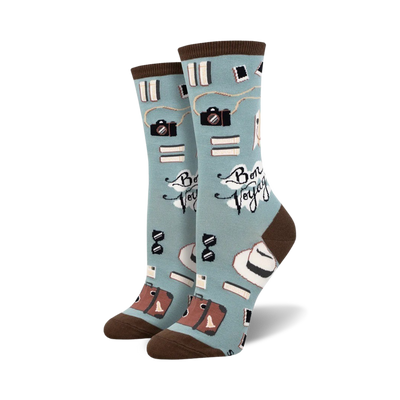 A pair of crew socks in teal and mint green, featuring cameras, books, hats, and suitcases; the phrase 'Bon Voyage' is printed on the socks.