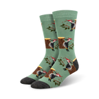 A pair of crew socks in sage green with a repeating pattern of woodpeckers on tree stumps and branches.