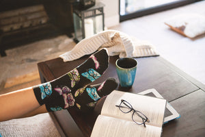 A person wearing black socks with a colorful pattern of eyeglasses on them is resting their feet on a coffee table. The table has a cup of coffee, a book, a tablet, and a pair of glasses on it. The person is relaxing in a living room with a fireplace and a large window in the background.