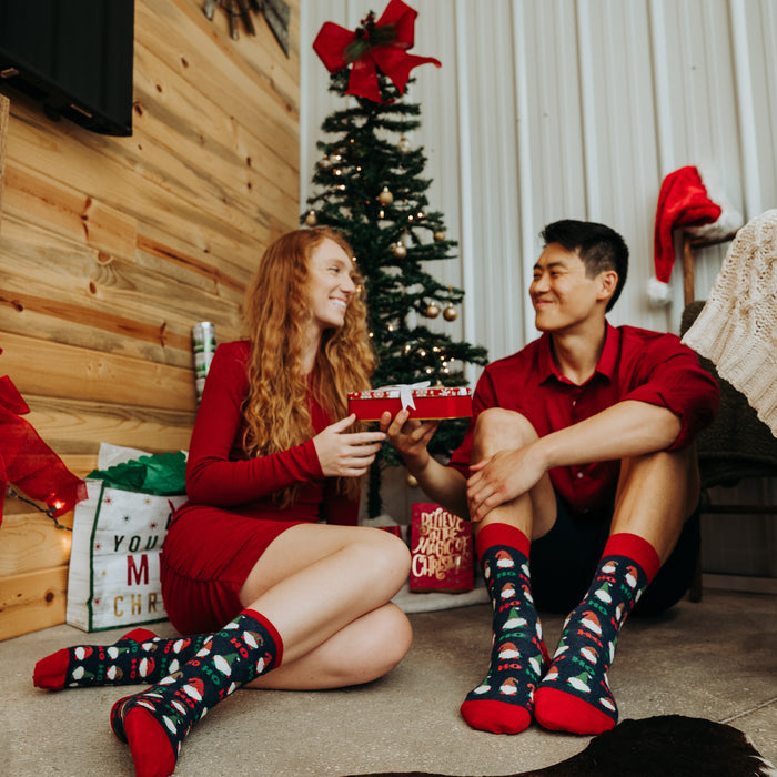 A man and woman are sitting on the floor in front of a Christmas tree. The woman is wearing a red dress and the man is wearing a red shirt and blue jeans. They are both wearing colorful socks and smiling at each other. The woman is holding a present.