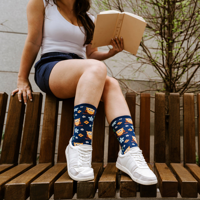A young woman is sitting on a park bench reading a book. She is wearing white sneakers, blue gym shorts, and colorful socks with an owl pattern.