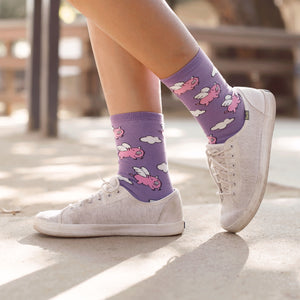A person is wearing a pair of purple socks with a pattern of pink pigs with wings on them. The person is also wearing a pair of white sneakers.