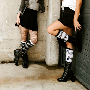 Two women are shown from the knees down. They are both wearing black skirts, black boots, and socks with a musical note pattern.