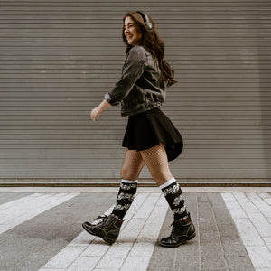 A young woman is walking down a crosswalk in the city. She is wearing a denim jacket, black skirt, fishnet stockings, black boots, and headphones. She has a smile on her face.
