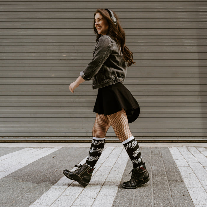 A young woman is walking down a crosswalk in the city. She is wearing a denim jacket, black skirt, fishnet stockings, black boots, and headphones. She has a smile on her face.
