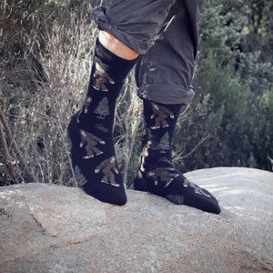 A man is putting on a pair of Darn Tough socks while sitting on a rock outdoors. He is wearing a pair of hiking boots and the socks are black with a pattern of pine trees and bears.