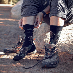 A man is putting on a pair of Darn Tough socks while sitting on a rock outdoors. He is wearing a pair of hiking boots and the socks are black with a pattern of pine trees and bears.