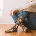 Model in jeans wearing brown iced coffee pattern crew sock with 'ICED. ALWAYS.' text on wood floor. [lf]