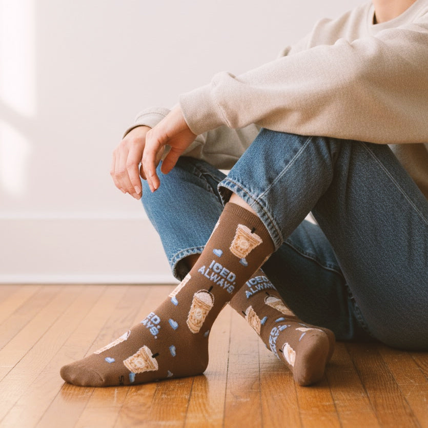 Model in jeans wearing brown iced coffee pattern crew sock with 'ICED. ALWAYS.' text on wood floor. [lf]