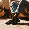 Person in cuffed jeans wearing black crew sock with golden acoustic guitar pattern, lounging indoors. [lf]