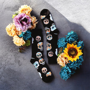 A pair of black socks with a pattern of sugar skulls on them. The socks are laying on a wooden table next to a bouquet of silk flowers in fall colors.