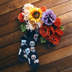 Overhead shot of Big Muertos Skull socks nestled amongst flowers on a wood background. [lf]