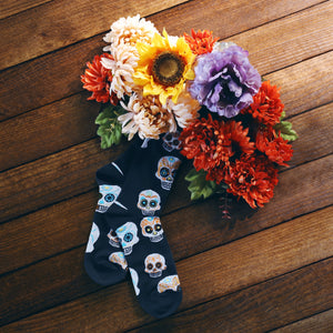 A pair of black socks with a pattern of sugar skulls on them. The socks are laying on a wooden table next to a bouquet of silk flowers in fall colors.