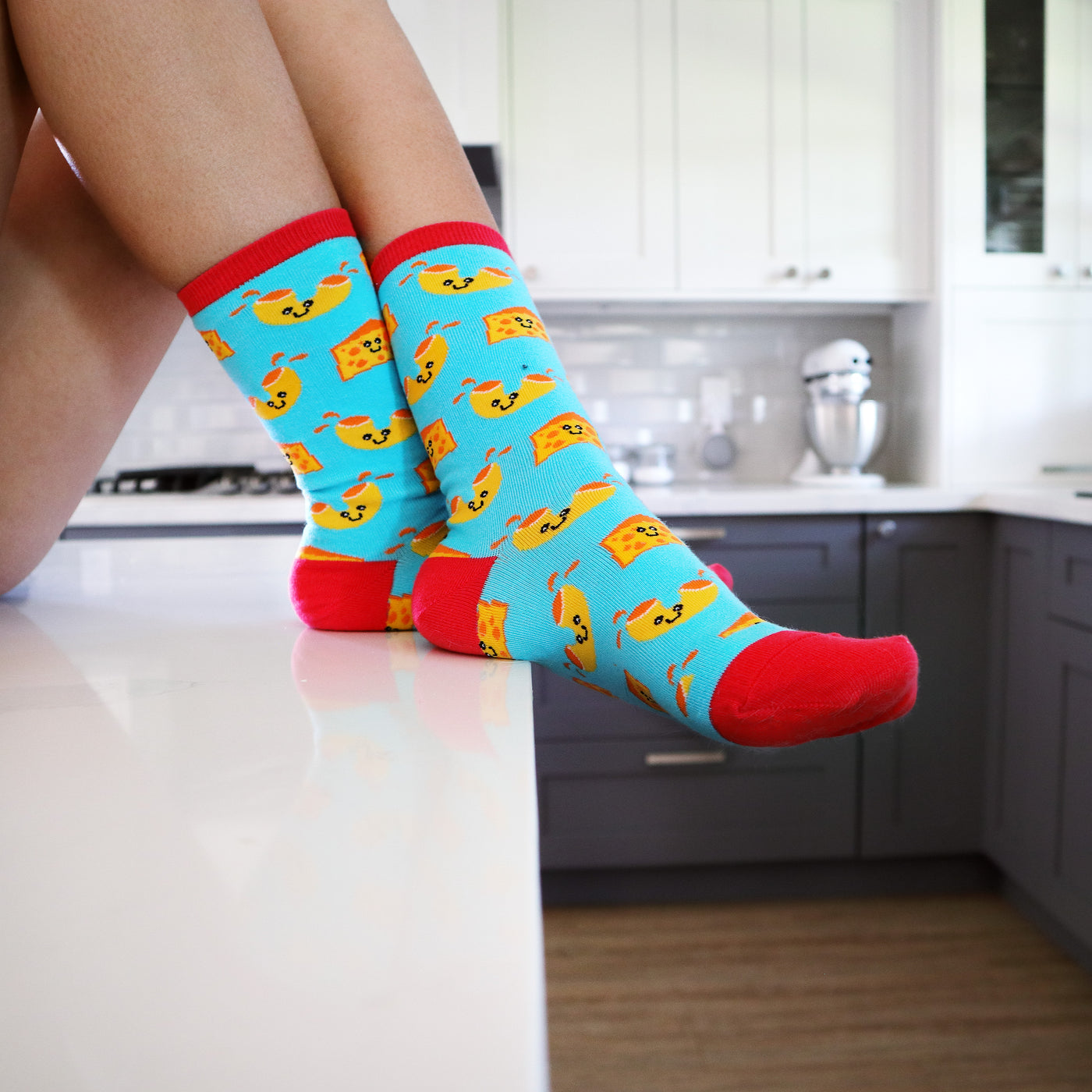 Lifestyle photo of Mac N Cheese socks on a person's legs, set in a kitchen. [lf]