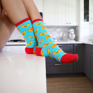 A person is sitting on a kitchen counter wearing blue socks with a pattern of cartoonish grilled cheese sandwiches. The tops of the socks are red.