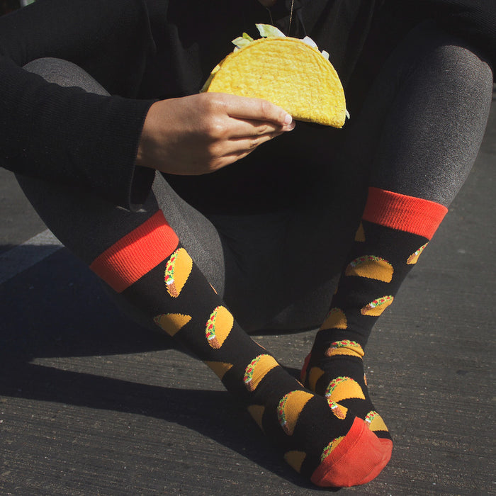 A person is holding a taco in their hand. The person is wearing black pants and socks with a taco pattern. The person is sitting on the ground.