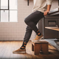 Model wearing black croissant crew socks with dark joggers and tan sneakers, leaning on a counter in a modern kitchen. [lf]