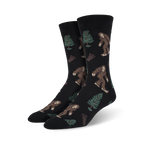 Pair of black Bigfoot XL crew socks. Features repeating pattern of Bigfoot, trees, and footprints. Brown, green, and black design on a black background.
