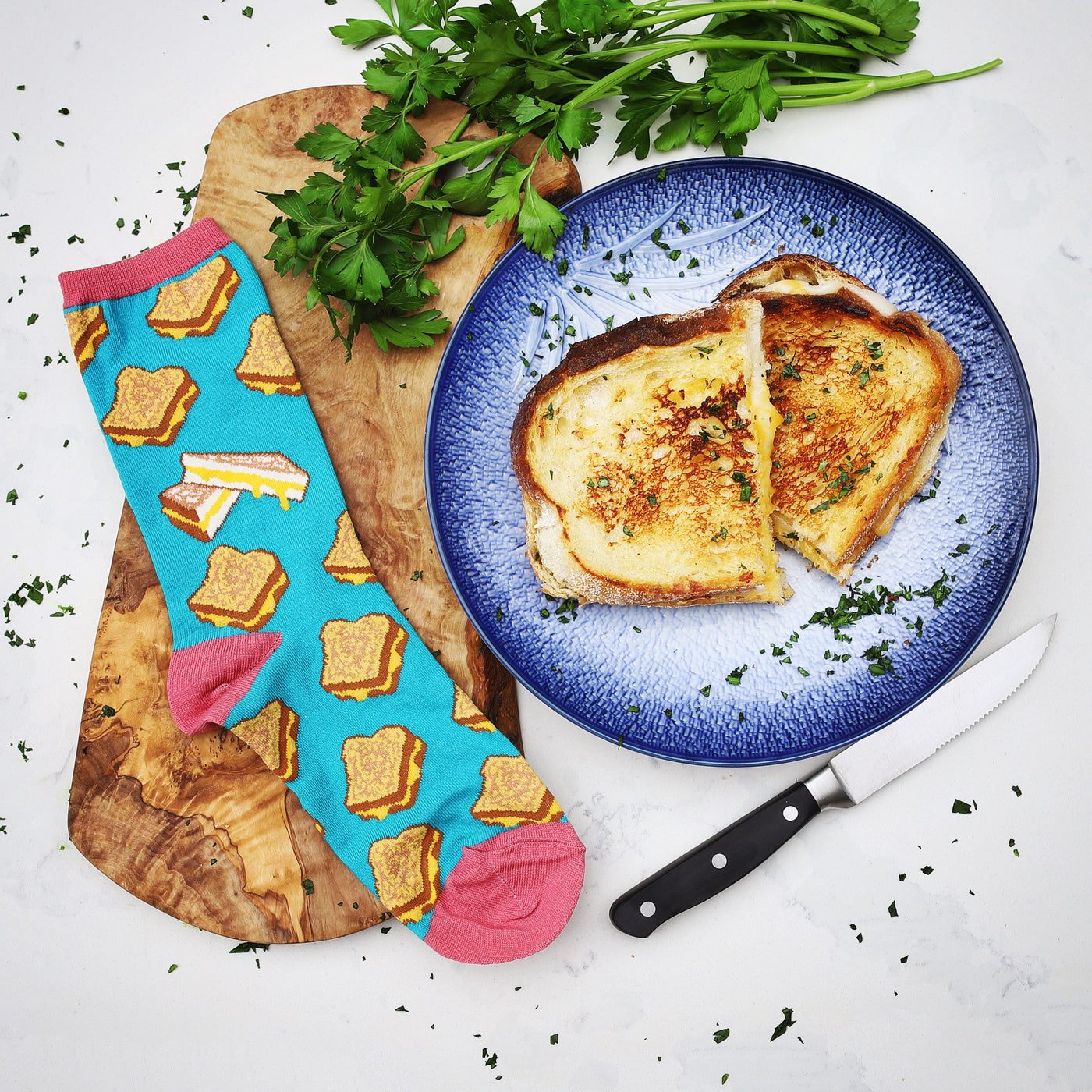 Lifestyle product shot of a single teal grilled cheese sock next to an actual grilled cheese sandwich on a plate. [lf]