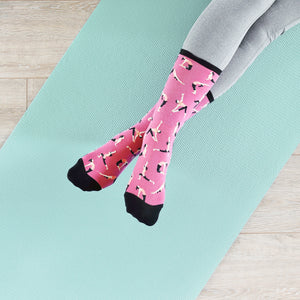 A person's feet in pink socks with a pattern of black yoga poses are resting on a blue yoga mat. The person is wearing gray sweatpants. The yoga mat is on a light brown wooden floor.