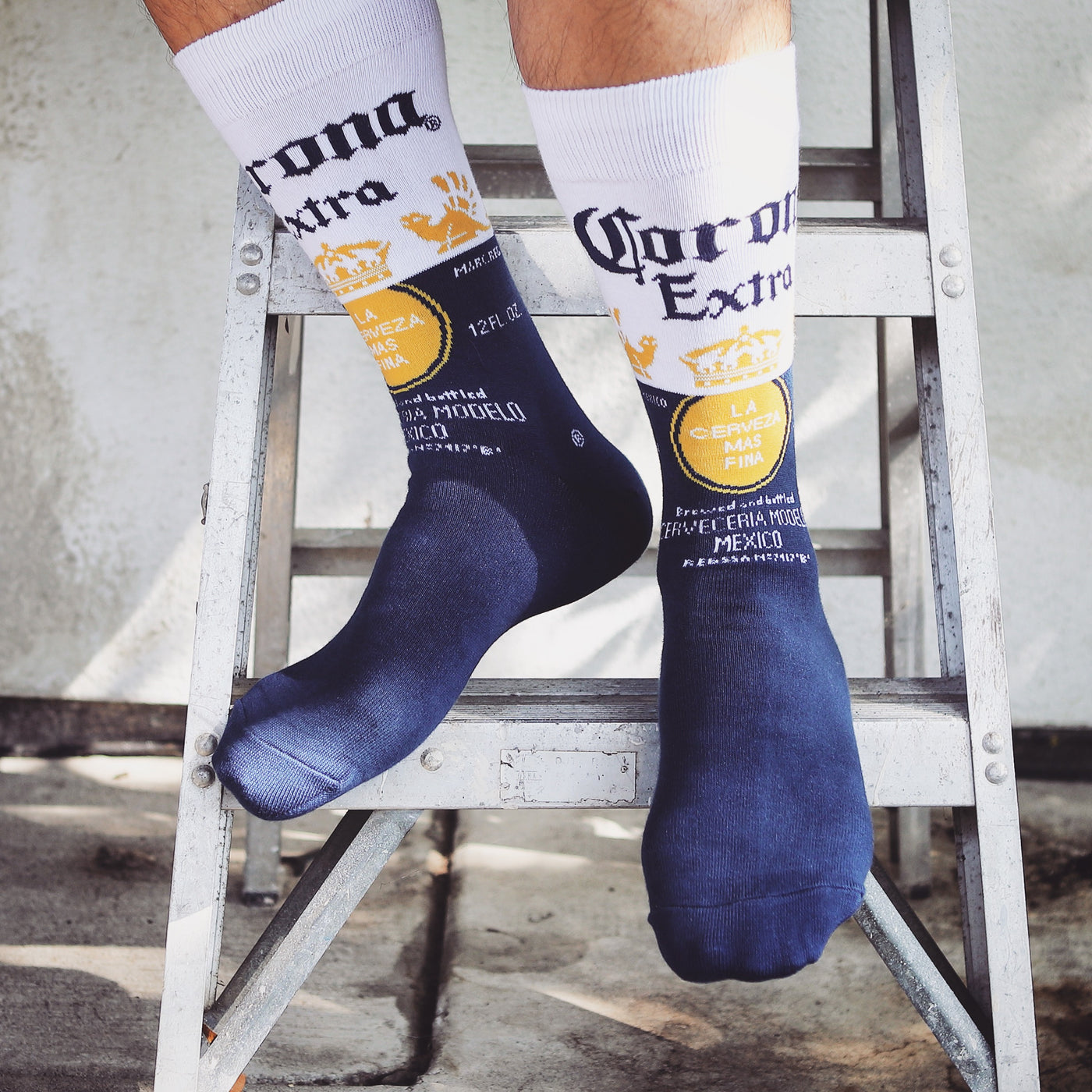 Lifestyle shot of a person's legs wearing the Corona Extra socks, sitting on a metal stepladder. [lf]