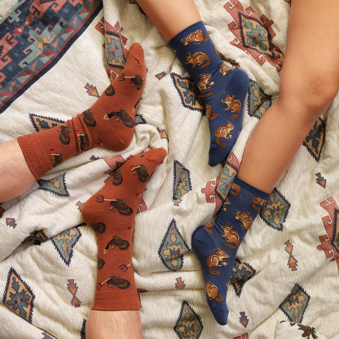 Two people are lying on a blanket with their feet up. One person is wearing brown socks with a pattern of turkeys holding forks. The other person is wearing blue socks with a pattern of chipmunks holding acorns.