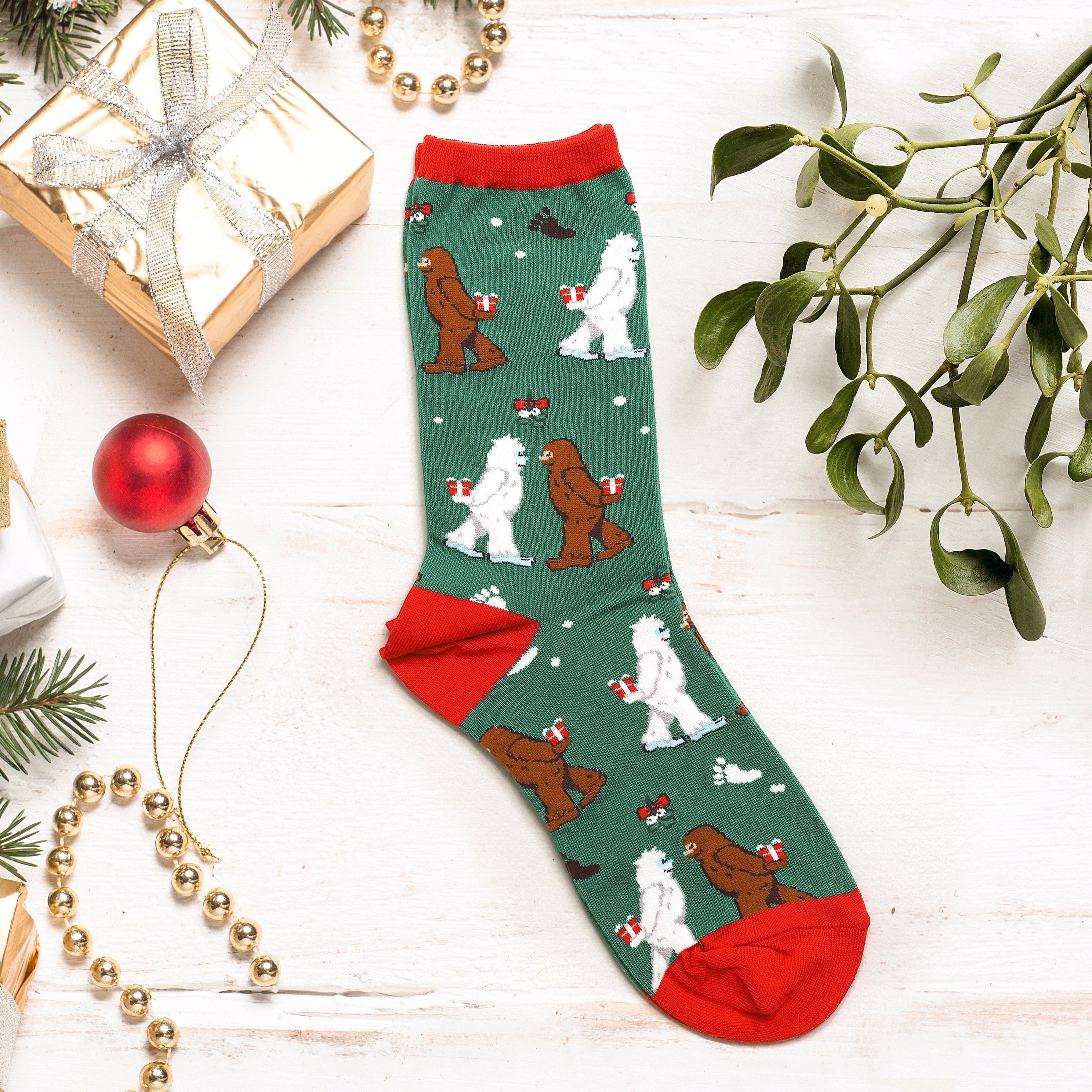 Flat lay of a green Mythical Kissmas sock against a white background with festive decorations. [lf]