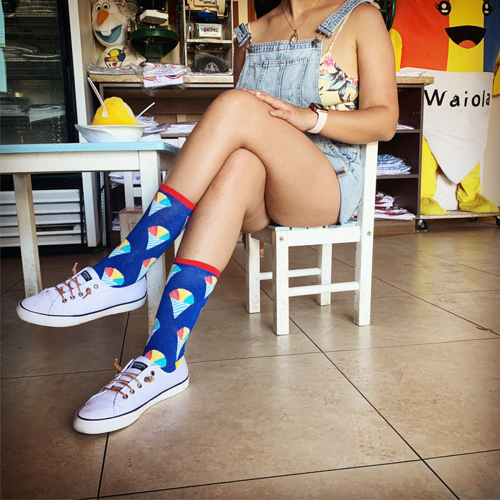 A woman is sitting on a white wooden chair with her legs crossed. She is wearing denim overall shorts, a white shirt, white sneakers, and colorful socks with an ice cream cone pattern. She is sitting at a table with a bowl of food on it. There are knick-knacks and a poster on the wall behind her.