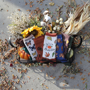 A black metal basket with a wooden handle is on the ground. The basket contains a variety of fall-themed socks, including socks with sunflowers, butterflies, and pumpkins. There are also some dried flowers, pine cones, and corn husks in the basket. The basket is sitting on a bed of brown and yellow fall leaves.