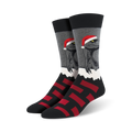 A pair of black, gray, and red crew socks. The main design is a velociraptor wearing a Santa hat, set against a snowy background. Red block pattern at the bottom.