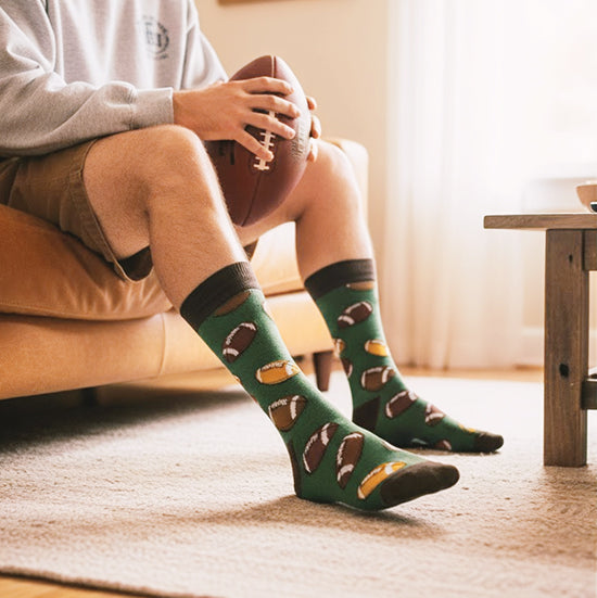 Green crew socks with scattered brown and tan footballs. Features a dark brown cuff, heel, and toe.