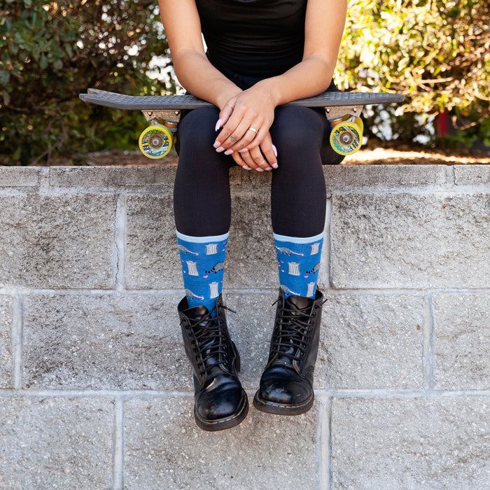 A young woman is sitting on a skateboard on a low brick wall outdoors. She is wearing black pants, black boots, and blue socks with a raccoon pattern. She has her hands resting on her knees.