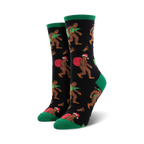 A pair of women's crew socks with a black background. Bigfoot characters wear Santa hats and carry Christmas trees. Green cuffs and red accents.