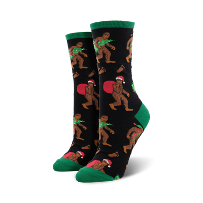 womens black crew socks with cartoonish brown bigfoot in red santa hats, carrying a christmas tree or sack of presents. green toes and cuffs.  