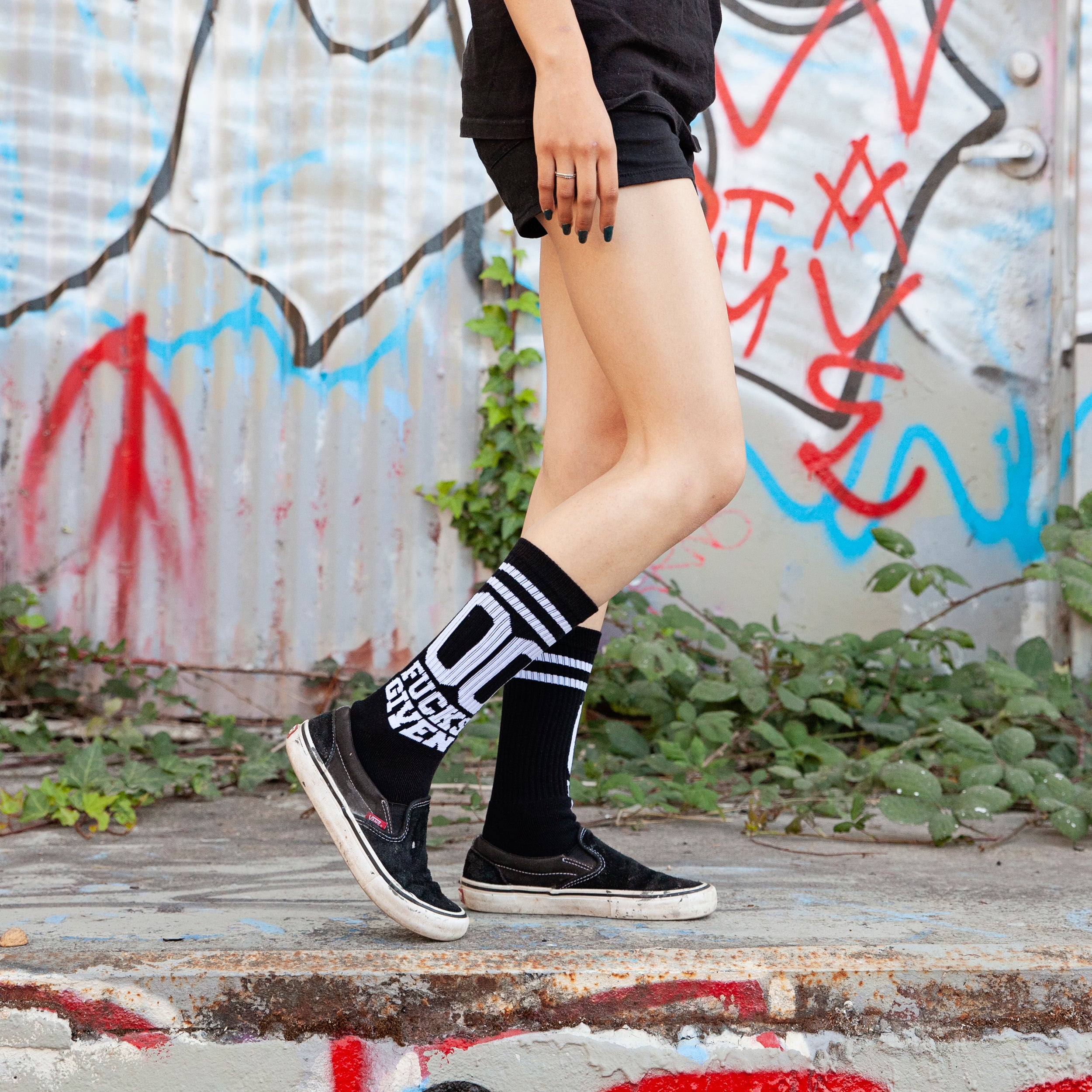 Lifestyle shot: Person wearing black '00 Fucks Given' socks with black Vans shoes against a graffitied wall. [lf]