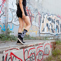 Lifestyle shot: Person wearing black '00 Fucks Given' socks and black shoes, sitting on a wall with graffiti background. [lf]